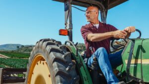 A man driving a tractor on a farm with fields and blue skies in the background.